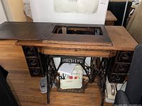 Full view of antique Singer sewing machine set in wooden cabinet with iron treadle base and knee pedal, showing storage drawers on sides.