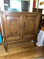 Front view of the wooden server cabinet showing two doors with decorative wood inlays and metal handles, standing on four square legs.