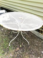 Full view of round white wrought iron table showing mesh top, central umbrella hole, and curved legs. Table is placed outdoors on dirt and grass against house siding.