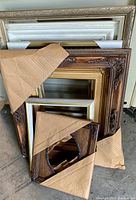 Photo showing multiple stacked mixed wood picture frames wrapped partially in brown paper. Frames include ornate dark wood, gold, white, and black with gold accents.