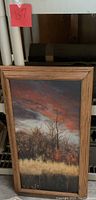 Vertical acrylic painting featuring an autumnal landscape with bare trees, a reddish cloudy sky, and yellow grass, framed in wood.