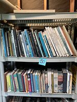 Two photographs showing two white plastic shelves filled with books and magazines. The first photo shows upper shelves with mostly hardcover books on various subjects including carpentry and regions. The second photo shows lower shelves with books and magazines, including more paperbacks and reference guides.