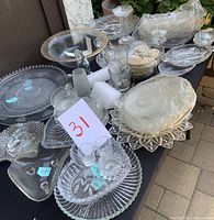 Photo showing arrangement of clear glass and crystal platters, bowls, water glasses, a candle holder, and a small rabbit decorative piece on a table outdoors.