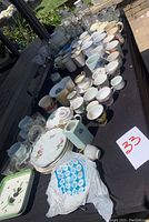 Overview of the lot showing assorted plates, cups, and glassware arranged on a black tablecloth outdoors.