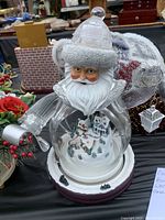 Clear Santa figurine with miniature winter village inside, white beard, and frosted hat brim and base sitting on black tablecloth with other Christmas decorations.