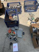 Overview of crates and boxes containing assorted electrical parts including metal covers, plugs, brackets, and other hardware spread on ground.