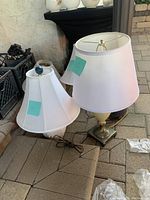 Two table lamps on the ground: one with a white fabric shade and hexagonal shape, blue rounded base; one with white conical fabric shade and marble base with brass accents