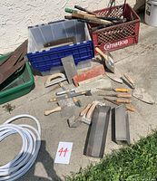 Overview of used hand tools and garden tools arranged on a pavement, showing metal and wooden handled saws, trowels, pruning clippers, and mixing containers.
