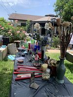 Outdoor photo showing long taper candles in various colors, a green ceramic pitcher vase holding artificial flowers, and other decorative items laid out on a table.