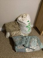 Silk mattress topper in white carrying bag and beige-colored bed linens folded next to a blue and gray patterned quilt with matching pillow shams.