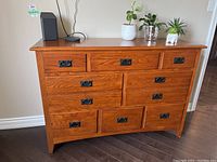 Front view of a wooden dresser with nine drawers, placed on a dark hardwood floor against a beige wall. Top surface has some plants and a speaker.