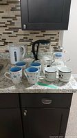 Set of four blue interior floral mugs, two black tree design mugs, two electric kettles (one clear glass, one white), glass teapot, and white ceramic holders.