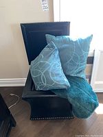 Black leather-like storage ottoman containing two decorative blue pillows with circular patterns and a matching blue plush throw blanket draped inside