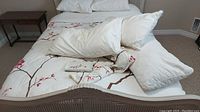 Bed made with white duvet cover featuring pink and brown floral branch pattern, three white pillows arranged on the bed, folded pillow covers placed on the bed, and white ceramic dish visible on the bedding.