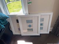 Three white wood-framed matching prints leaning against a wall near a window.