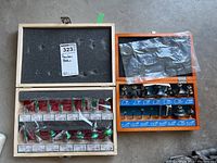 Photo showing two boxes of router bits side by side on a concrete floor, one wooden box open revealing neatly arranged labeled green-topped router bits, the other an orange plastic box containing assorted bits.