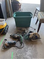 Power tools on garage floor including Black and Decker circular saw, Hitachi drill, Bosch jig saw, and green plastic tote bin