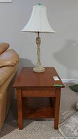 Side view of wood side table with shelf and table lamp on top near a beige sofa.