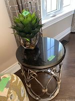 Round side table with black marble-like top and metal base, displaying the faux plant on top near window.