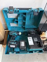 Open blue plastic carrying case with Makita cordless drill, battery charger, and additional battery compartment.