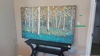 Full view of the canvas wall art showing birch trees in a forest with blue and gold colors, resting above a black table.