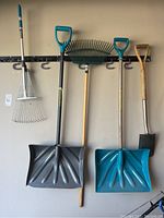 Photo showing five yard tools hanging on a garage wall including two large snow shovels, two metal leaf rakes with blue handles, one green plastic garden rake, and a wooden-handled metal spade.