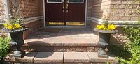 Two identical black cast iron urn planters with soil and yellow flowering plants on brick steps