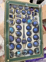Photo showing a green box with a set of blue and gold Christmas ball ornaments, including round, decorated, and finial shapes, some metallic and matte finishes.