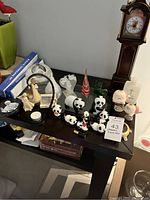 Wide angle view of shelf displaying ceramic panda figurines, Bowring tall clock, baptismal candles, glass oil lamp, and other small ceramic figures