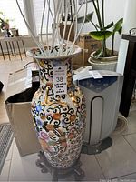 Photo of tall ceramic Chinese vase with colorful intricate floral design and wooden stand beneath it. Vase has a small chip and is 24 inches tall without stand.