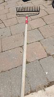 Garden soil rake with wooden handle and black metal tines on brick pavement.