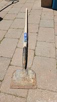 Large metal spade shovel with wooden handle, showing rust and wear, laying on outdoor pavers.