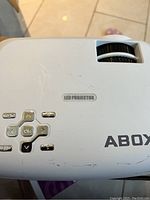 Close-up photo of the top of the Abox LED projector showing the LED projector label, the manual focus wheel, some scratches, and partial brand name 'ABOX'.