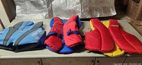 Three life jackets laid out on a table: blue Speedo vest, red Coleman vest, red/yellow Buoy-O-Boy vest with visible adjustable straps and foam padding.