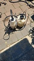 Two white plastic hand-pump garden sprayers with hoses placed on stone patio