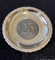 Front view of 25th anniversary silver commemorative plate showing embossed 25th emblem and ornate border.