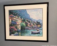 Framed artwork showing a waterfront village with boats, buildings, hills, and a blue sky.