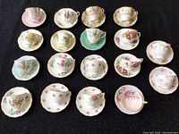 Top view of entire set showing 18 assorted tea cups and 18 saucers arranged in rows on dark fabric background with various floral designs.