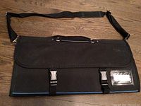 Front view of the closed black chef's carrying bag showing flap with buckles, carry handle, shoulder strap, and ID window.