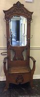 Front view of antique wooden hall seat with carved details, mirror, metal hooks on sides for hanging items, armrests and umbrella holder rings on sides.