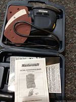 Top view of Mastercraft detail sander/polisher in molded blue plastic carrying case with electrical cord coiled, multiple triangular sanding discs, and instruction manual visible.