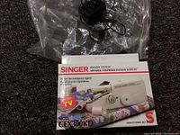 Front view of the original box showing Singer Handy Stitch sewing machine and included power adapter, cables, and manual.