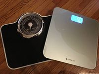 Mechanical and digital bathroom weight scales side by side on wooden floor showing condition and display