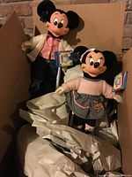 Two vintage Mickey and Minnie Mouse dolls standing on white doll stands inside a cardboard box, with packing paper around.