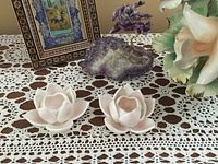 Two lotus-shaped ceramic candle holders, white with pink tint, placed near a polished stone and a flower figurine on a crocheted tablecloth
