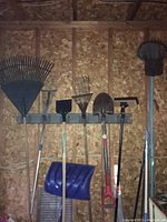 Photo of seven gardening tools hanging on a wall including rakes, hoes, shovels, and a push broom