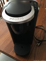 Front and top view of black and silver Keurig coffee maker on wooden table with visible power cord.