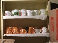 Shelf with two rows of vintage cups including milk glass cups on top row and copper tone cups on bottom row