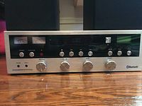 Front view of silver Bluetooth mini audio system showing control knobs, power button, function selector, CD tray, digital display, and Bluetooth logo with two speakers on sides.