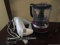 Photo showing KitchenAid mini food chopper and Sunbeam hand mixer side by side on a brown surface.